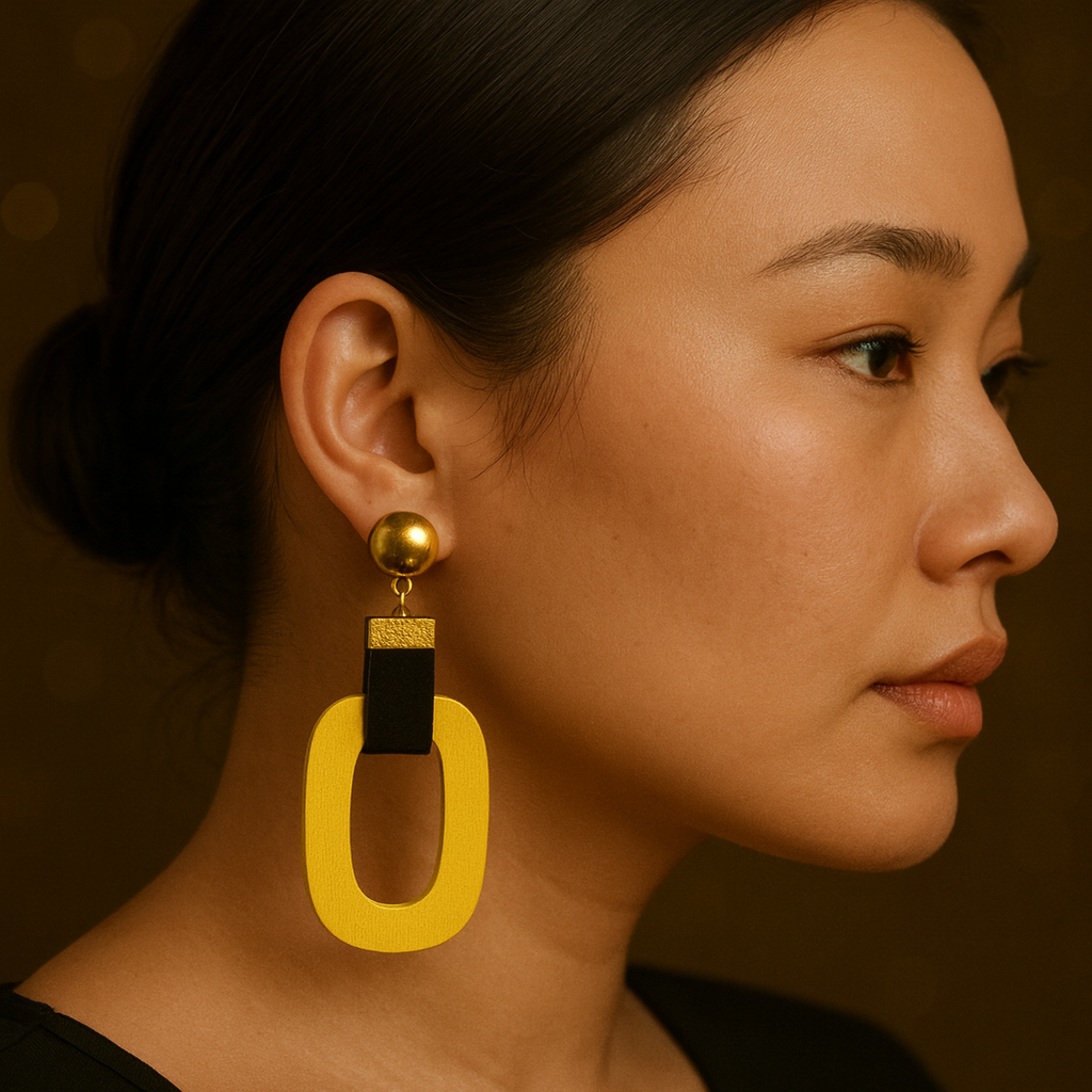 A close-up profile of a young East Asian woman wearing a bold statement earring with a gold stud, black velvet connector, and a large yellow oval-shaped pendant.