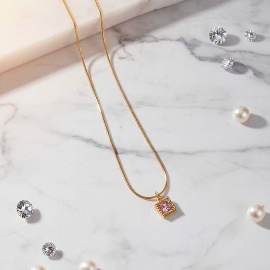 Gold necklace with a square pendant on a marble surface with scattered pearls and crystals.