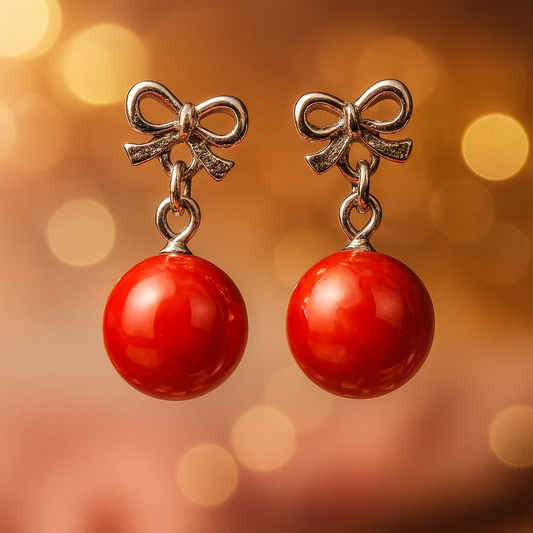 Pair of silver bow earrings with glossy red spherical drops, displayed against a warm bokeh background.