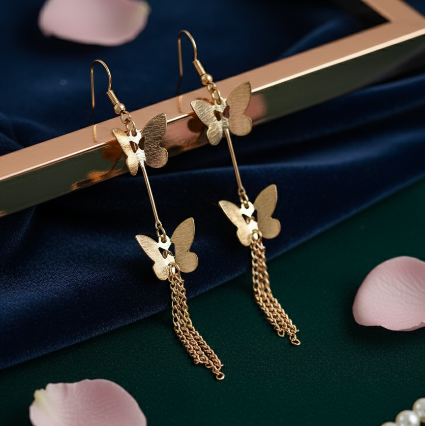 Gold butterfly earrings with chains on a dark surface with pink petals