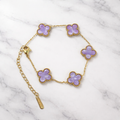 A timeless bracelet crafted in a delicate gold-tone finish, adorned with dreamy lilac clover charms on a white marble background. 