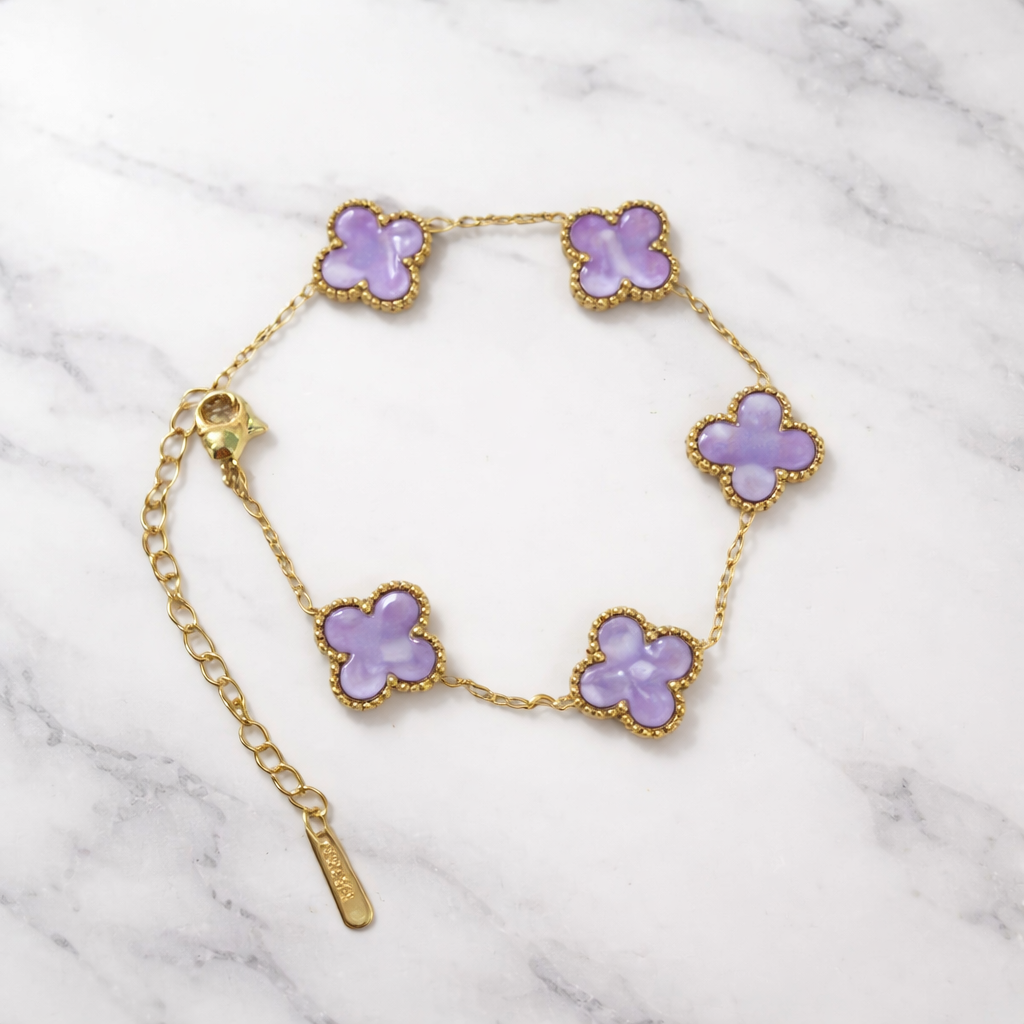 A timeless bracelet crafted in a delicate gold-tone finish, adorned with dreamy lilac clover charms on a white marble background. 