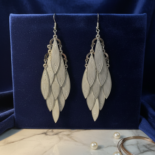 Pair of silver leaf-shaped earrings on a blue cushion with pearls and bangles in the background.
