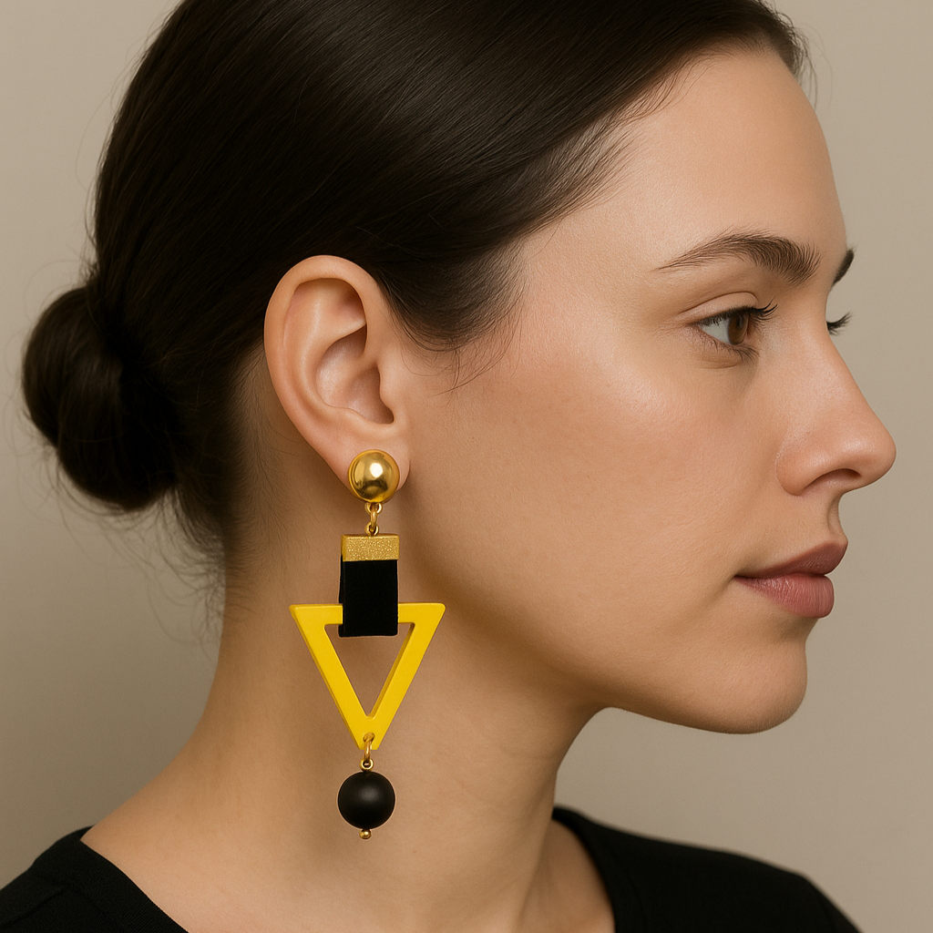 A close-up profile of a woman with fair skin and tied-back dark hair wearing a bold geometric statement earring featuring a gold stud, black velvet strap, yellow inverted triangle frame, and a dangling black bead.