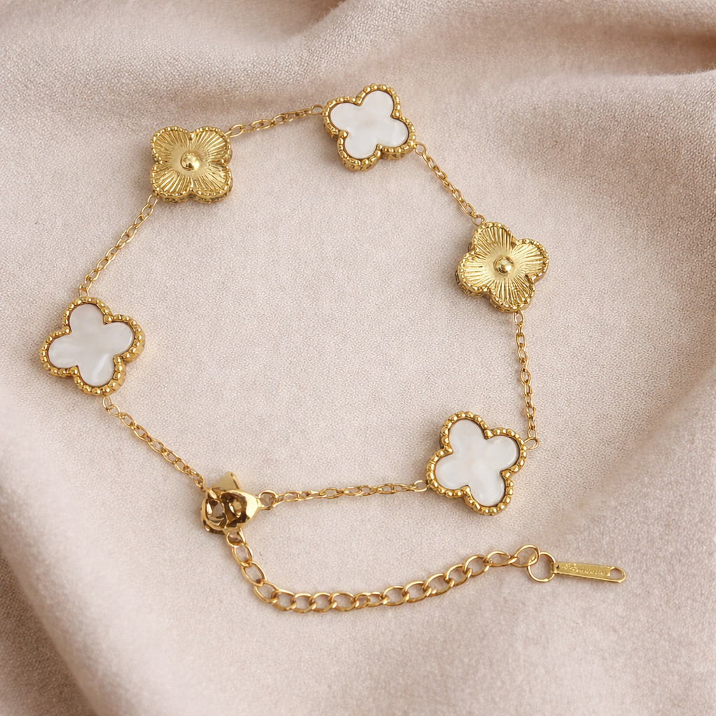 A golden bracelet with textured gold floral motifs and white mother-of-pearl clovers framed with beaded gold edges on a champagne cloth. 